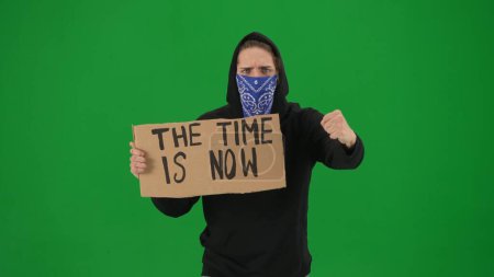 Foto de Un manifestante masculino con la cara cubierta por una bufanda sostiene un letrero que dice que el tiempo es ahora. El hombre en una pantalla verde de cerca. Clave de croma - Imagen libre de derechos