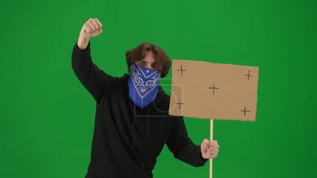 Téléchargez les photos : Militant masculin avec le visage couvert d'écharpe, criant des slogans de protestation et tenant un panneau vierge. Un manifestant sur l'écran vert se rapproche. Publicité, promo, clé chromatique - en image libre de droit