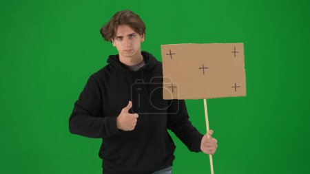Photo for A male activist holds a blank poster and shows a thumbs up. A young man in casual clothes holds a placard on a green screen in close up. Chromakey, advertisement, promo - Royalty Free Image