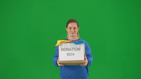 Photo for A woman volunteer holds a box full of clothes labeled Donation box. Portrait of female volunteer holding donation box on green screen close up. Concept of social assistance, volunteering, humanitarian - Royalty Free Image