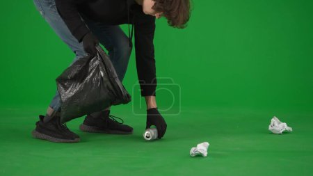 Téléchargez les photos : Un volontaire homme portant des gants noirs et portant un sac poubelle ramasse les ordures sur l'écran vert de près. Aide volontaire au travail gratuit concept d'aide - en image libre de droit