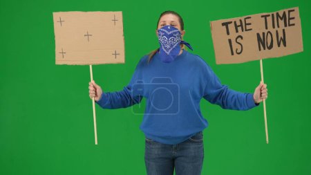 Foto de Una manifestante con la cara cubierta por una bufanda sostiene un cartel que dice que el tiempo es ahora y un signo en blanco. La mujer activista en una pantalla verde de cerca. Clave de croma - Imagen libre de derechos
