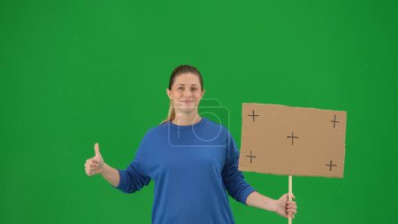 Photo for Female demonstrator holding a blank poster and showing a thumbs up. A young woman in casual clothes holding a poster on a green screen close up. Chroma key, advertisement, promo - Royalty Free Image