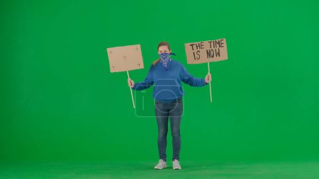 Photo for A female demonstrator with her face covered by a scarf holds up a sign that says Time is now and a blank sign. The woman activist on a green screen. Chroma key - Royalty Free Image