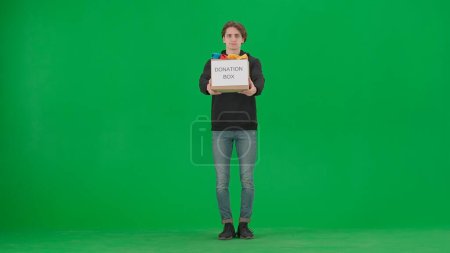 Photo for A man volunteer holds a box full of clothes labeled Donation box. The man holds out the box on the green screen. Concept of social assistance, volunteering, humanitarian and charity. Helping people - Royalty Free Image