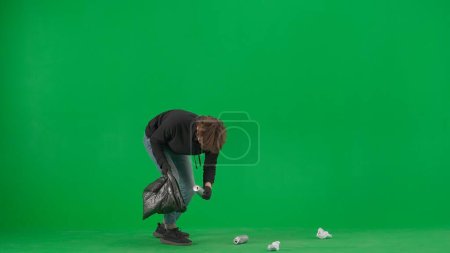 Foto de Un voluntario masculino que lleva guantes negros y una bolsa de basura recoge la basura en la pantalla verde. El hombre pone papel y latas en la bolsa de basura. Concepto de ayuda voluntaria para el trabajo gratuito - Imagen libre de derechos