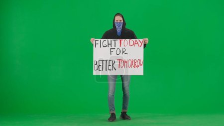 Photo for A male activist holds a placard that reads Fight today for a better tomorrow. A man in a hoodie with his face covered by a scarf on a green screen. Protest. Motivation. Rally. Future. Fighting. Rally - Royalty Free Image