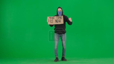 Foto de Un manifestante masculino con la cara cubierta por una bufanda sostiene un letrero que dice que el tiempo es ahora. El hombre manifestante en una pantalla verde. Clave de croma - Imagen libre de derechos