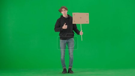 Photo for A male activist holds a blank poster and shows a thumbs up. A young man in casual clothes holds a placard on a green screen. Chromakey, advertisement, promo - Royalty Free Image