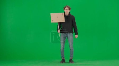 Téléchargez les photos : Un militant masculin triste tient une affiche vierge sur un écran vert. Clé chromatique, publicité, promo - en image libre de droit