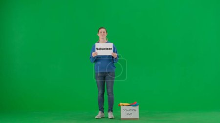 Téléchargez les photos : Une femme tenant une affiche avec le mot Bénévole dessus et une boîte de vêtements à ses pieds avec le mot Boîte de don dessus. Volontaire féminine en studio sur écran vert. Aider les gens - en image libre de droit