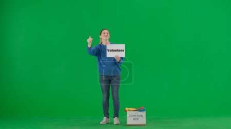 Foto de Una mujer voluntaria agita la mano, mostrando el gesto de venir aquí en el estudio en la pantalla verde. Mujer sosteniendo un cartel con la inscripción Voluntariado, una caja de ropa a sus pies etiquetada Donación - Imagen libre de derechos