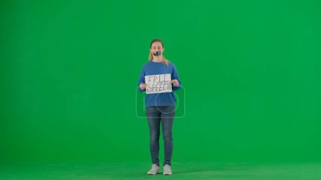 Photo for A woman with her mouth taped with black tape holds a poster with the inscription Free speech. Protestant woman fighting for her rights on a green screen. Conceptualizing the idea of free speech and - Royalty Free Image