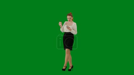 Foto de Mujer de negocios moderna. Retrato de atractiva chica de oficina en croma pantalla verde clave. Mujer en falda y blusa caminando bebiendo café de la taza, mensajes de texto en el teléfono inteligente. Media vuelta.. - Imagen libre de derechos