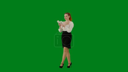 Téléchargez les photos : Femme d'affaires moderne. Portrait d'une jolie fille de bureau sur écran vert chroma. Femme en jupe et chemisier marchant et comptant l'argent, expression joyeuse du visage. Demi-tour. - en image libre de droit