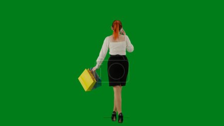 Téléchargez les photos : Femme d'affaires moderne. Portrait d'une jolie fille de bureau sur écran vert chroma. Femme en jupe et chemisier marchant parlant sur smartphone. Vue arrière. - en image libre de droit