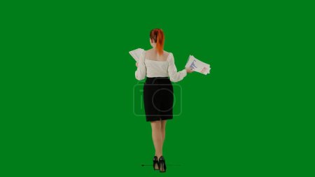 Téléchargez les photos : Femme d'affaires moderne. Portrait d'une jolie fille de bureau sur écran vert chroma. Femme en jupe et chemisier marchant lisant des documents en papier. Vue arrière. - en image libre de droit