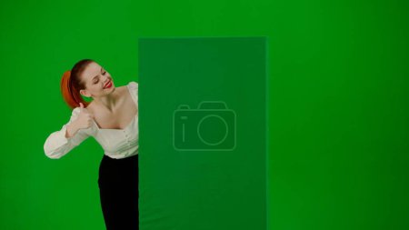 Foto de Retrato de atractiva chica de oficina en pantalla verde con cromakey. Mujer en una falda y blusa mira hacia fuera desde detrás de un cartel publicitario, sonríe y señala un pulgar hacia arriba. - Imagen libre de derechos