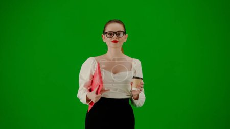 Foto de Mujer de negocios moderna concepto de publicidad creativa. Retrato de atractiva chica de oficina en croma pantalla verde clave. Mujer en falda y gafas caminando sosteniendo carpeta roja y taza de café, cara sonriente. - Imagen libre de derechos