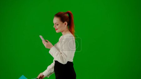 Téléchargez les photos : Femme d'affaires moderne concept de publicité créative. Portrait d'une jolie fille de bureau sur écran vert chroma. Femme en jupe marchant tenant des sacs-cadeaux et envoyant des SMS sur smartphone. Vue latérale. - en image libre de droit