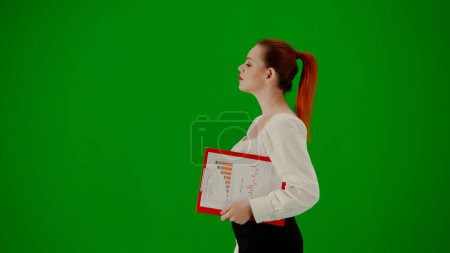 Foto de Mujer de negocios moderna concepto de publicidad creativa. Retrato de atractiva chica de oficina en croma pantalla verde clave. Mujer en falda y blusa caminando sosteniendo carpeta roja. Vista lateral. - Imagen libre de derechos