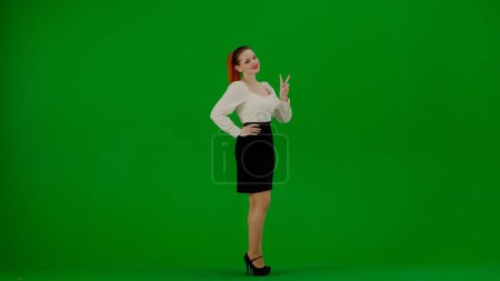 Foto de Mujer de negocios moderna concepto de publicidad creativa. Retrato de atractiva chica de oficina en croma pantalla verde clave. Mujer en falda y blusa de pie sonriendo a la cámara mostrando signo de victoria. - Imagen libre de derechos