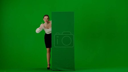 Photo for Portrait of attractive office girl on green screen with chromakey. Woman in a skirt and blouse looks out from behind a advertising billboard, smiles and points a thumbs up. - Royalty Free Image