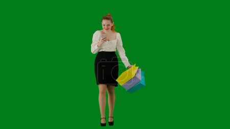 Foto de Mujer de negocios moderna concepto de publicidad creativa. Retrato de atractiva chica de oficina en croma pantalla verde clave. Mujer en falda caminando sosteniendo bolsas de regalo y mensajes de texto en el teléfono inteligente, cara sonriente. - Imagen libre de derechos
