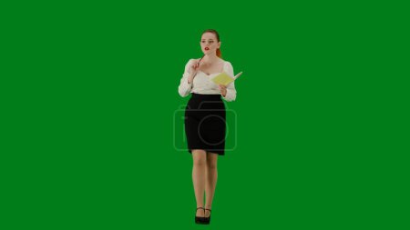 Téléchargez les photos : Femme d'affaires moderne concept de publicité créative. Portrait d'une jolie fille de bureau sur écran vert chroma. Femme en jupe et chemisier marchant tenant un cahier et écrivant des idées, visage concentré. - en image libre de droit
