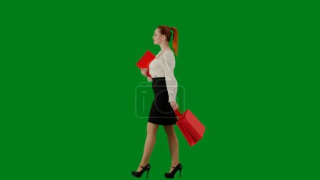 Foto de Mujer de negocios moderna. Retrato de atractiva chica de oficina en croma pantalla verde clave. Mujer en falda y blusa caminando con carpeta de papel y bolsas de papel de regalo rojo. Vista lateral. - Imagen libre de derechos