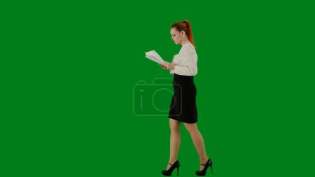 Téléchargez les photos : Femme d'affaires moderne. Portrait d'une jolie fille de bureau sur écran vert chroma. Femme en jupe et chemisier marchant en lisant des documents en papier, expression focalisée du visage. Vue latérale. - en image libre de droit