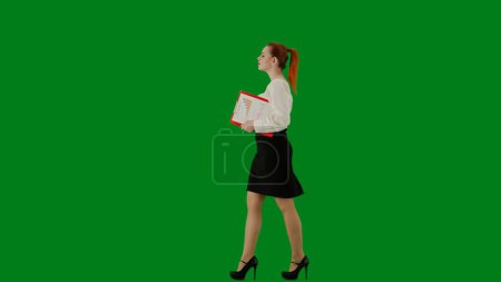 Téléchargez les photos : Femme d'affaires moderne concept de publicité créative. Portrait d'une jolie fille de bureau sur écran vert chroma. Femme en jupe et chemisier marchant tenant dossier rouge. Vue latérale. - en image libre de droit
