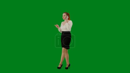 Téléchargez les photos : Femme d'affaires moderne. Portrait d'une jolie fille de bureau sur écran vert chroma. Femme en jupe et chemisier marchant parlant sur smartphone, expression positive du visage. Demi-tour. - en image libre de droit
