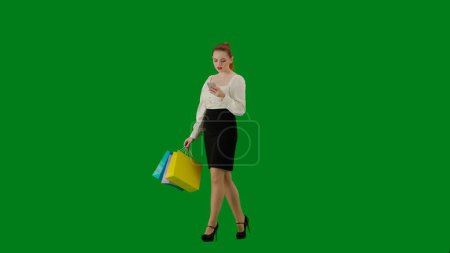 Foto de Mujer de negocios moderna concepto de publicidad creativa. Retrato de atractiva chica de oficina en croma pantalla verde clave. Mujer en falda caminando sosteniendo bolsas de regalo y mensajes de texto en el teléfono inteligente. Media vuelta.. - Imagen libre de derechos
