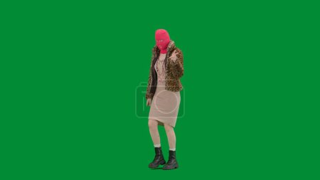 Foto de Mujer con pasamontañas rosa, abrigo de tigre y vestido apuntando con el dedo índice en dirección. Mujer loca sobre fondo verde en el estudio. Concepto de tendencias de la moda, tendencia feminista en la moda. Media vuelta. - Imagen libre de derechos