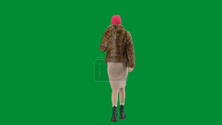 Foto de Mujer en pasamontañas rosa, abrigo de piel de tigre y vestido de noche caminando. Mujer freak en fondo verde en el estudio. Concepto de tendencia de moda, tendencia feminista en la moda. Vista trasera - Imagen libre de derechos