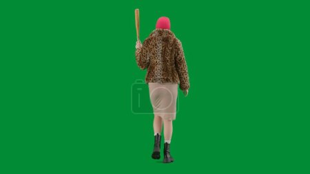 Téléchargez les photos : Femme en cagoule rose, manteau en fourrure de tigre et robe de soirée marchant avec une batte à la main. Femme monstre sur fond vert en studio. Vue arrière - en image libre de droit