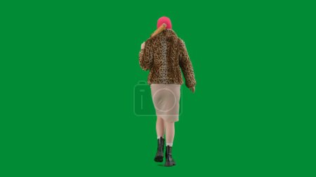 Téléchargez les photos : Femme en cagoule rose, manteau en fourrure de tigre et robe de soirée marchant avec une batte à la main. Femme monstre sur fond vert en studio. Vue arrière - en image libre de droit