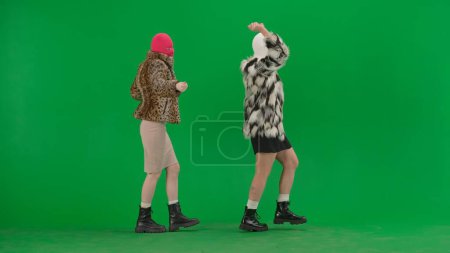 Foto de Dos mujeres en pasamontañas blancas y rosadas caminando y bailando alegremente. Mujeres raras en abrigos de piel sobre fondo de estudio verde. Concepto de tendencia de moda, tendencia feminista en la moda - Imagen libre de derechos