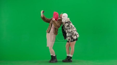 Photo for Two women in white and pink balaclavas taking selfies using smartphone. Freak women in fur coats on green background studio. Fashion trend concept, feminist trend in fashion - Royalty Free Image