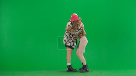 Photo for Two women in white and pink balaclavas looking expectantly at a smartphone and rejoicing at good news or victory. Freak women in fur coats on green studio background. Fashion trend concept. - Royalty Free Image