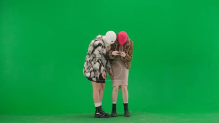 Foto de Dos mujeres en pasamontañas blancas y rosadas jugando un juego en un teléfono inteligente. Mujeres raras en abrigos de piel en estudio de fondo verde. Concepto de tendencia de moda, tendencia feminista en la moda - Imagen libre de derechos