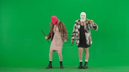 Photo for Two women in white and pink balaclavas dancing funny with a bat and a crowbar. Freak women in fur coats on green studio background. Fashion trend concept, feminist trend in fashion - Royalty Free Image