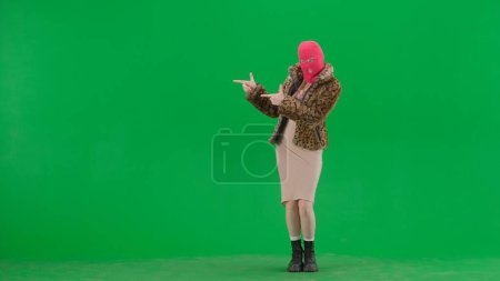 Téléchargez les photos : Femme en cagoule rose, manteau de tigre et robe de soirée pointe les doigts sur le côté. Une femme bizarre sur fond vert dans un studio - en image libre de droit