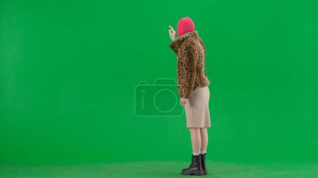 Foto de Mujer en pasamontañas rosa, abrigo de tigre y el grifo vestido de noche, deslizar en la pantalla virtual. Mujer loca sobre fondo verde en el estudio. Concepto de tendencia de moda, tendencia feminista en la moda - Imagen libre de derechos