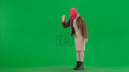 Téléchargez les photos : Femme en cagoule rose, manteau de tigre et robinet de robe de soirée, glisser sur l'écran virtuel. Femme bizarre sur fond vert en studio. Concept de tendance de la mode, tendance féministe dans la mode - en image libre de droit