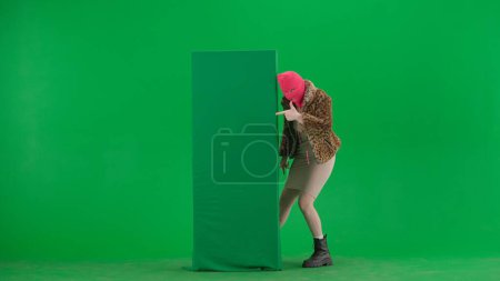 Téléchargez les photos : Femme en cagoule rose, manteau de tigre et robe pointant du doigt la bannière verte. Une femme bizarre sur fond vert dans le studio - en image libre de droit