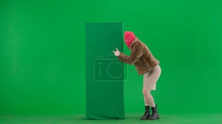 Téléchargez les photos : Femme en cagoule rose, manteau de tigre et robe pointant du doigt la bannière verte. Une femme bizarre sur fond vert dans le studio - en image libre de droit
