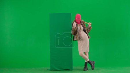 Foto de Mujer en pasamontañas rosa, abrigo de tigre y vestido apoyado en la bandera verde y haciendo gesto de dedo ir aquí. Una mujer rara con un bate en sus manos sobre un fondo verde en un estudio. Concepto tendencia moda - Imagen libre de derechos