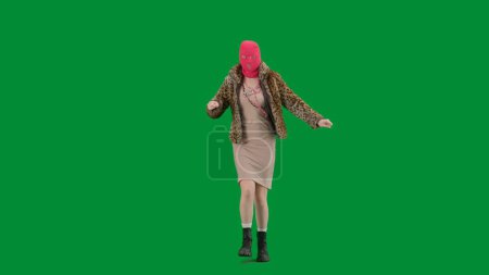 Foto de Mujer en pasamontañas rosa, abrigo de piel de tigre y vestido de noche bailando alegremente. Mujer freak en fondo verde en el estudio. Concepto de tendencia de moda, tendencia feminista en la moda - Imagen libre de derechos